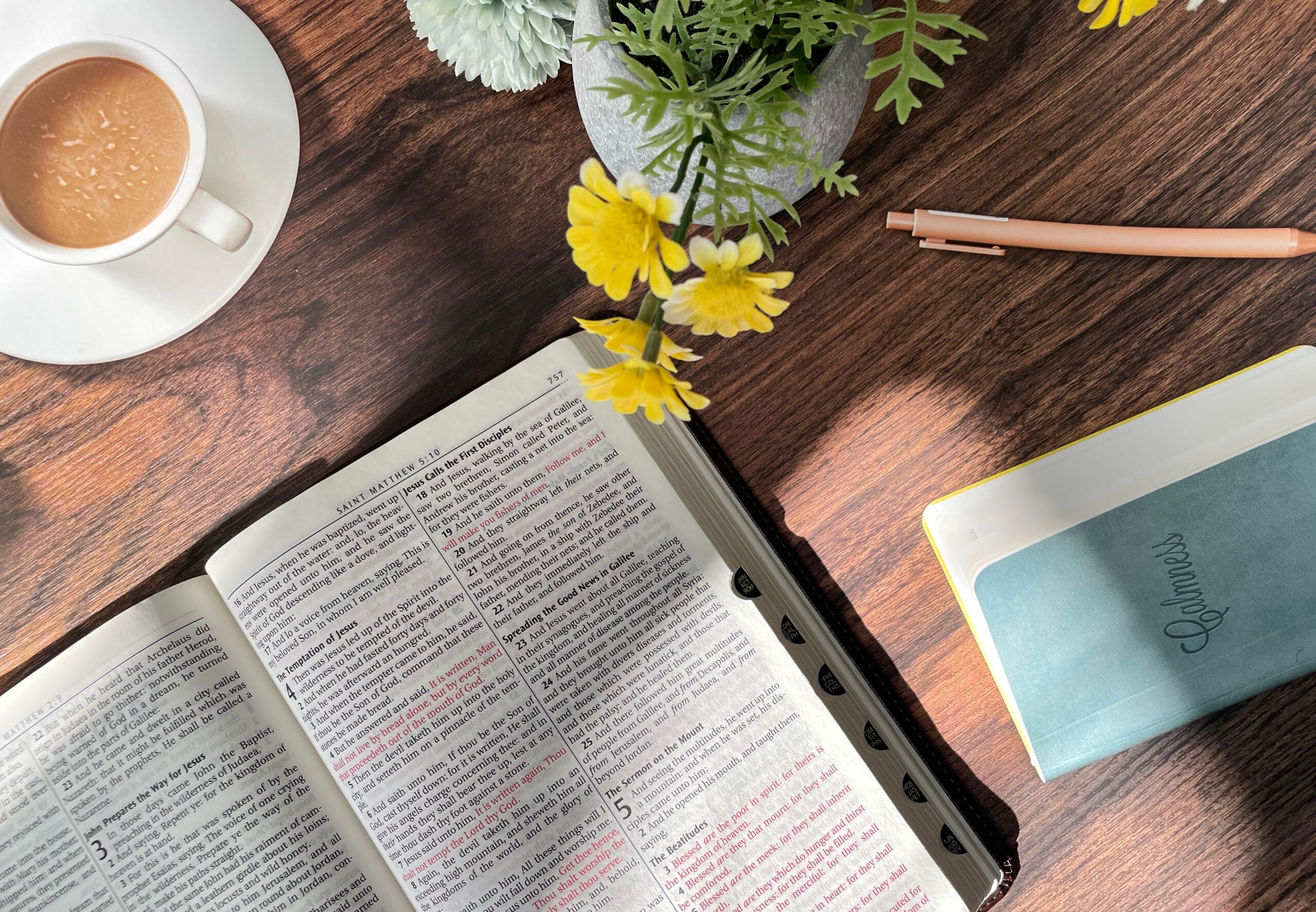 Flat lay showing an open Bible, coffee cup, notebook, and flowers on a wooden table in sunlight.